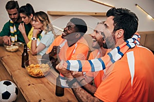 side view of happy young multicultural friends in orange fan t-shirts celebrating goal in soccer match while their friends