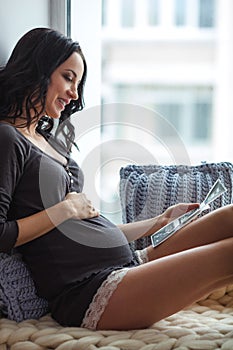 Side view of happy young pregnant woman looking at ultrasound scan on window sill