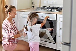 Side view of happy young woman and cute kid daughter wearing casual attires bake dessert in kitchen, teaching child making