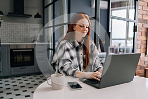 Side view of happy young woman typing on laptop computer sitting at table with coffee cup smiling looking to screen in