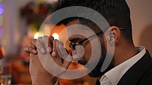 Side view headshot of young Middle Eastern man in eyeglasses praying sitting indoors on Christmas night. Close-up