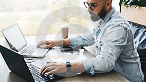 Side view. Hipster bearded businessman sits in cafe, works on two laptops. Freelancer work remotely. Online education