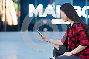 Side view of hipster woman in trendy outfit using mobile phone sitting outside of shopping mall
