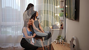 A side view of how three girls, friends, look at themselves in a backlit mirror, straighten their hair and smile, while