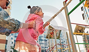 Side view image of active cute little girl climbing on a rope at playground. Mother helping her daughter to climb on a wall with a