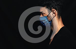 Side view image of Caucasianman wearing medical blue mask on the face during virus pandemic lockdown posing on the black wall with