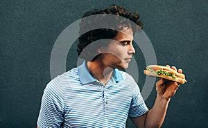 Side view image of young handsome man eating a healthy burger. Hungry man in a fast food restaurant eating a hamburger outdoors.