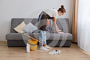 Side view image of young woman cleaning her home, wearing jeans and brown apron, housewife doing her household chores at home in