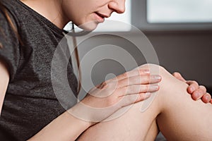 Side view of irritated young woman sitting on bed at home massaging touching knee with hands, feeling pain.