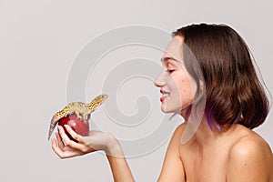 Side view. Isolation. Inside. Young and brunette girl holding apple on which sits an gecko.