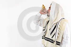 Side view of a Jewish man in tallith blowing the Shofar against a white background.