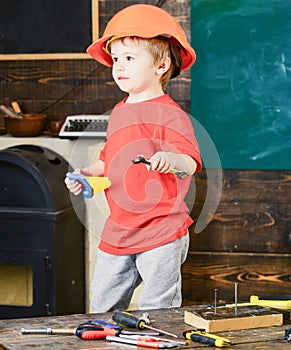 Side view kid standing in workshop. Blond boy in orange helmet holding spanner and handsaw. Little repairman posing for