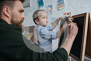 side view of little boy and father with pieces of chalk drawing picture on blackboard