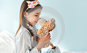 Side view of a little girl wearing pajama in stripes blue and white, smiling broadly, playing with toy giraffe in the bed,