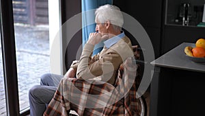 Side view of lonely senior man in wheelchair thinking looking out floor-to-ceiling window. Thoughtful Caucasian male