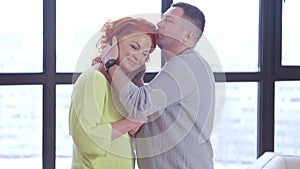 Side view of loving adult man kissing and hugging woman standing with spouse at window indoors. Happy confident husband