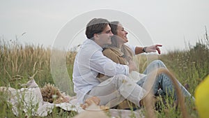 Side view loving man hugging woman sitting on picnic pointing away talking. Smiling Caucasian girlfriend enjoying date