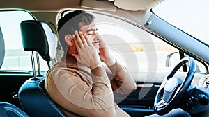 Side view male driver clutches his head with his hands and grimaces from the pain in his head.