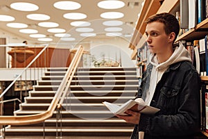 Side view of male student in library holding a book. University student standing at bookshelf