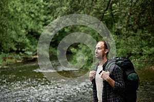 Side view of a man with a large hiking backpack standing on the bank of the river and enjoying nature