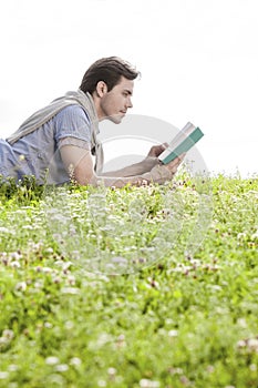 Side view of man reading book while lying on grass against clear sky
