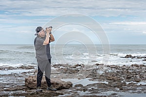 Side view of mature adult photographer with cap taking a picture focusing towards the sky on the rocks near the sea.