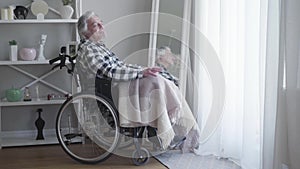 Side view of mature Caucasian grey-haired man sitting in wheelchair and thinking. Lonely elderly man spending day alone