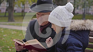 Side view of a mature Caucasian man in classic clothes sitting on the bench with his granddaughter and reading a book in