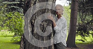 Side view of mature woman hugging tree and smiling outdoor.