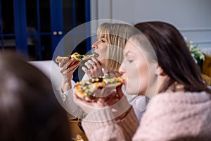 Side view of middle-aged women friends relatives sit at table, holding pieces of pizza, eating, enjoying food at home.