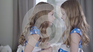 Side view middle shot of angry little Caucasian girl scolding her twin sister. Pretty siblings in blue dotted dresses