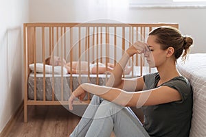 Side view of mother with postnatal depression symptoms sitting on floor while baby sleeping in bed