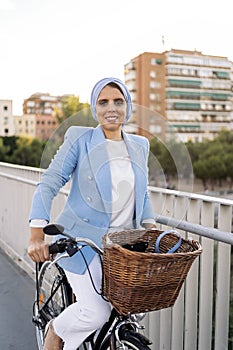 Side view of a muslim woman standing with her bike on a footbridge looking at camera