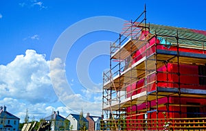 Side view of a new two-storey house under construction with red protective layer, scaffolding and wooden boards.