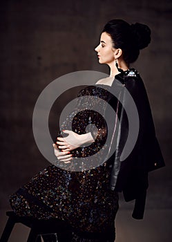 Side view of noble pregnant noble woman in black off-shoulder dress and leather jacket stands holding hand at her belly