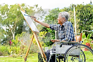 Side view of one Asian senior man sit on wheelchair and paint the painting outdoor for relax and revive good health of old people