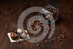 Side view of overturned glass jar with coffee beans and chocolate candies on wooden background, selective focus