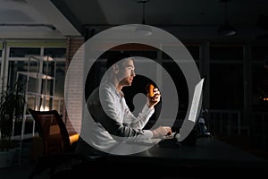 Side view of overworked businessman eating burger working on computer sitting on desk at office workplace late night in