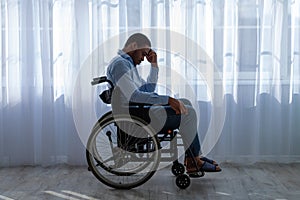 Side view of paraplegic black man feeling desperate and lonely, having depression, sitting in wheelchair near window