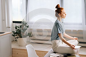 Side view of peaceful redhead young woman is meditating while sitting on desk at home office.