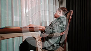 Side view of a person sitting at a desk with a thoughtful pose, looking away from the computer, with curtains in the background