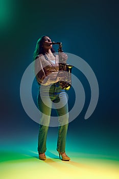 Side view photo of Indian woman, saxophonist performing solo in neon light against gradient background.