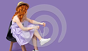 Side view photo of a redhead young pretty woman in sundress and strae hat holds shoelaces in hands of her sneekers