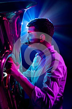 Side view photo of talented man, artistic musician seated, playing trumpet with bright blue-pink stage lights behind him