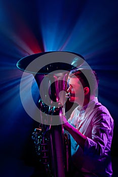 Side view photo of talented man, artistic musician seated, playing trumpet with soft blue-pink stage lights behind him.