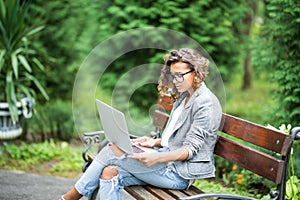 Side view of pleased brunette woman in eyeglasses sitting on bench in park and using laptop computer