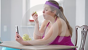 Side view of plus-size Caucasian young woman eating healthful organic vitamin apple looking at smartphone screen