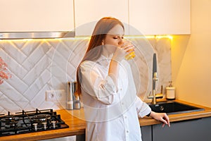 Side view portrait of attractive young woman in casual clothes drinking orange juice from glass standing in kitchen.