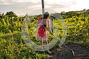 Side view portrait of beautiful teen girl with brunette lng hair wering red dress working in vineyard with rakes