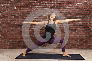 Side view portrait of beautiful young woman wearing black top and purple leggings working out against brick wall, doing yoga or